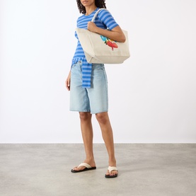 Model wearing Ganni Butterfly Logo Striped T-Shirt in Nouvean Navy, styled with denim shorts, carrying a cream tote bag, wearing flip flops.
