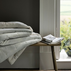 Christys platinum folded duvet displayed on a wooden bench beside an open book and green plant near a window