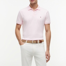 Model wearing Tommy Hilfiger Pink Lily polo shirt with short sleeves and logo detail, styled with white trousers and tan belt.