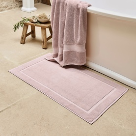 Christys Petal Pink bath mat with border detail, styled in a bathroom setting with a matching towel draped over a freestanding bath.