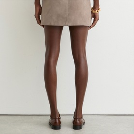 Model wearing Reiss beige mini skirt, styled with brown loafers and gold chain bracelet, rear view