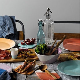 Royal Doulton multi-coloured dinnerware set styled on a rustic table with food, a glass soda siphon, wooden board and linen napkins.