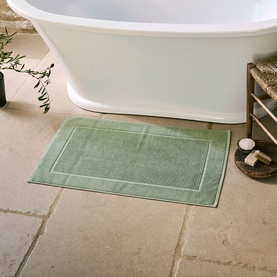 Christys Bay Leaf Green bath mat with double border detail, styled on stone floor beside a freestanding white bathtub