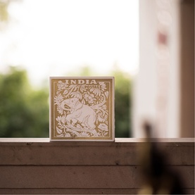 Archivist India candle in decorative kraft box with intricate animal and floral stamp-style print, displayed on wooden ledge outdoors