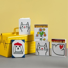 Archivist matchbox set with illustrated cat, bird, bear and floral designs, arranged in front of a yellow tin box