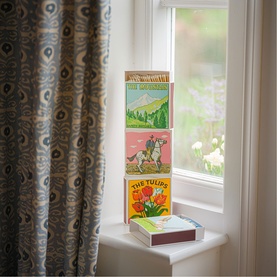 Archivist gift box set stacked on a windowsill, featuring vintage-style illustrated labels including The Tulips, beside a patterned curtain