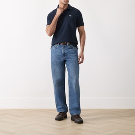 Model wearing Barbour navy cotton polo shirt, styled with mid-wash straight-leg jeans and a brown leather belt.