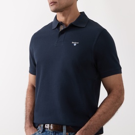 Model wearing Barbour navy cotton polo shirt with contrast collar and embroidered chest logo, styled with blue jeans and brown belt.