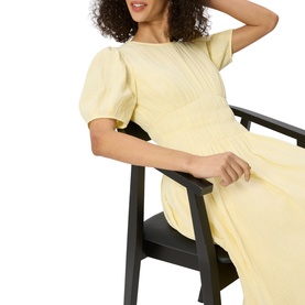 Model wearing Whistles yellow dress with puff sleeves and gathered midi skirt, seated on a dark wooden chair