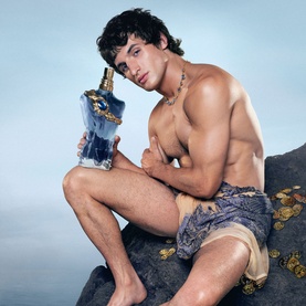 Model with Jean Paul Gaultier Le Beau Narcisse Eau de Parfum, shirtless seated on rocks holding the blue and gold bottle