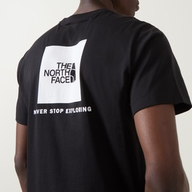 Model wearing North Face Evolution Box Logo Back Graphic T-Shirt in black, showing white square logo and "Never Stop Exploring" text on back.