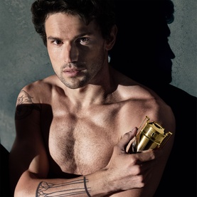 Model with Paco Rabanne Invictus Elixir Parfum, gold bottle held against chest, shirtless male model with tattoos