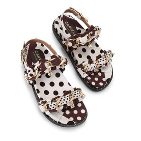 White and brown polka dot flat sandals with gold chain and floral embellishment detail, ankle strap, and dark rubber sole.