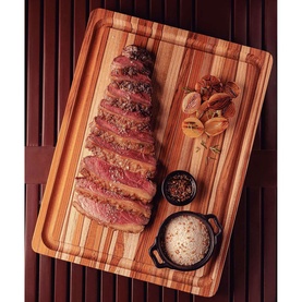 Tramontina brown wooden chopping board styled with sliced steak, roasted garlic, peppercorns, and a small dish of salt