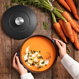 Tramontina black casserole dish with lid, filled with orange soup garnished with herbs, styled on wooden surface with fresh carrots