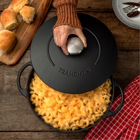Tramontina black cast iron casserole dish filled with mac and cheese, lid being lifted, styled with bread rolls on wooden board