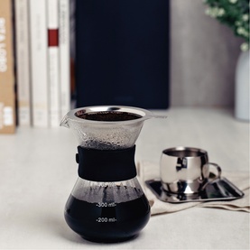 Glass pour-over coffee maker with black silicone sleeve and stainless steel filter, shown with espresso cups on a marble surface