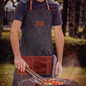 Model wearing Tramontina cotton and leather apron in dark grey, using grilling tongs over an outdoor barbecue.