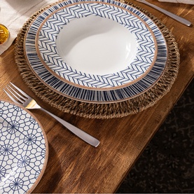 Tramontina blue and white geometric patterned dinner set with gold-tone rim detail, styled on a wooden table with woven charger plate and fork