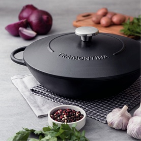 Tramontina black cast iron casserole dish with silver knob lid, styled with garlic, red onions and peppercorns on grey surface