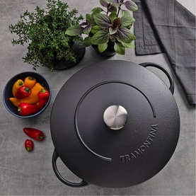Tramontina black cast iron casserole dish with lid and silver knob, styled on grey surface with fresh herbs and vegetables nearby.