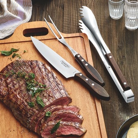 Tramontina black carving knife, fork, and tongs with dark wood handles on wooden board beside sliced grilled steak