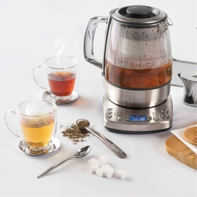 Stainless steel and glass electric tea maker with digital display, shown with brewed tea in glass cups, loose leaf tea and sugar cubes