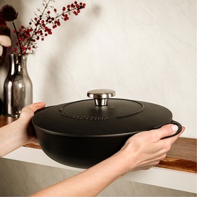 Tramontina black cast iron lidded casserole dish with silver knob handle, held by hands over wooden surface with vase in background