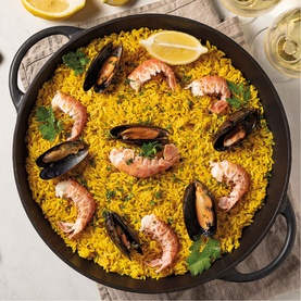 Tramontina black paella dish filled with saffron rice, prawns, mussels, and parsley, styled with lemon wedges and white wine glasses