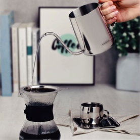 Tramontina stainless steel pour-over kettle in use, pouring water into a glass coffee dripper, with a small double-wall cup nearby