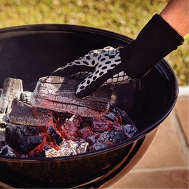 Tramontina black heat-resistant barbecue mitt worn while arranging hot coals and grill grate on a charcoal kettle BBQ