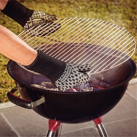 Tramontina black heat-resistant BBQ mitt worn while placing grill grate onto charcoal kettle barbecue outdoors