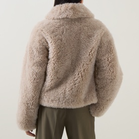 Model wearing Yves Salomon beige shearling coat, rear view, with khaki trousers. Fluffy textured finish, relaxed fit.