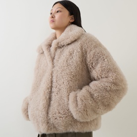 Model wearing Yves Salomon beige shearling coat with fluffy texture, spread collar, and relaxed silhouette, styled with dark trousers.