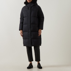 Model wearing Yves Salomon black longline puffer jacket with faux fur-trimmed hood, styled with dark cropped trousers and black loafers.