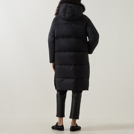 Model wearing Yves Salomon black longline puffer coat with fur-trim hood, shown from behind, styled with black trousers and flat shoes.