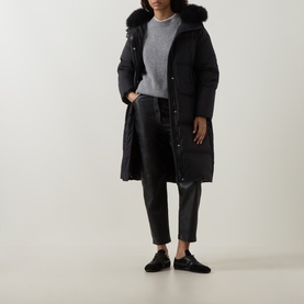 Model wearing Yves Salomon black long padded coat with fur-trimmed hood, styled with grey knit, cropped leather trousers and black trainers.