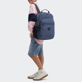 Model carrying Kipling Seoul Lap Backpack in Game Grey, with two front zip pockets, worn on back, styled with denim shorts and pink sweatshirt.
