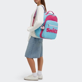 Model carrying Kipling Seoul Lap Backpack in Smile Blue Bl with pink Smile lettering, worn with white jumper and denim midi skirt.