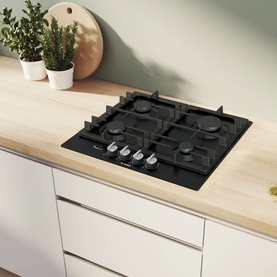 Bosch black four-burner gas hob set into a light wood worktop in a white kitchen, with silver knobs and cast iron pan stands.