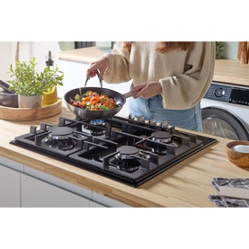 Bosch black gas hob set into wooden worktop, shown in use with a frying pan of vegetables being stirred by a model