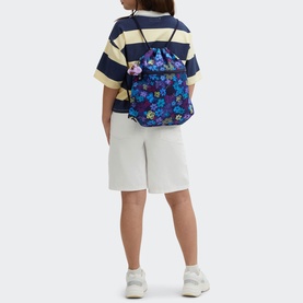 Model carrying Kipling Supertaboo Gym Bag in Dark Flwr Stamp, blue floral drawstring backpack, styled with yellow striped tee and white shorts