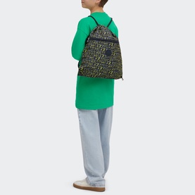 Model carrying Kipling Supertaboo Gym Bag in Lets Play Prt print, black with neon yellow lettering, drawstring straps, styled with green top and grey