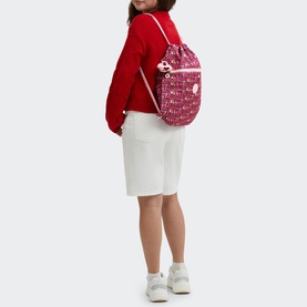 Model carrying Kipling Supertaboo Gym Bag in U R Great Prt red print, worn as backpack with drawstring straps, styled with red top and white shorts.