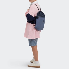 Model carrying Kipling Supertaboo Gym Bag in Game Grey, worn as backpack with drawstring straps, styled with pink coat and denim shorts