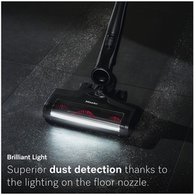 Miele cordless stick vacuum in black with illuminated floor nozzle, shown on dark tiled floor highlighting dust detection lighting feature
