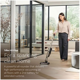 Model using Miele Triflex HX3 Plus cordless stick vacuum in champagne, vacuuming a bright open-plan living room with wood flooring
