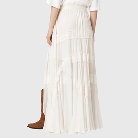 Model wearing All Saints Dorina Asymmetric Skirt in antique white with tiered lace panels and high-low hem, styled with a white tee and tan boots.