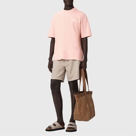 Model wearing All Saints Odyssey Oversized Bubble Logo T-Shirt in Blushed Pink, styled with beige shorts, brown sandals, and a tan tote bag.