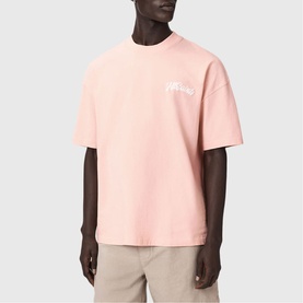 Model wearing All Saints Odyssey Oversized Bubble Logo T-Shirt in Blushed Pink with white bubble logo print, styled with beige trousers.